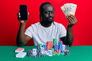 handsome young black man playing poker holding smartphone dollars skeptic nervous frowning upset because problem negative person